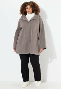 Cappotto grigio fluffoso e oversize con cappuccio, caratterizzato da una chiusura a bottoni e maniche ampie, abbinato a pantaloni neri e sneakers bianche.