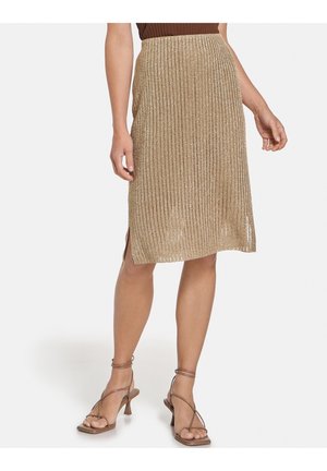 Femme portant une jupe midi plissée métallisée dorée avec une fente sur le côté et des sandales à talons marron à brides, debout devant un fond blanc.