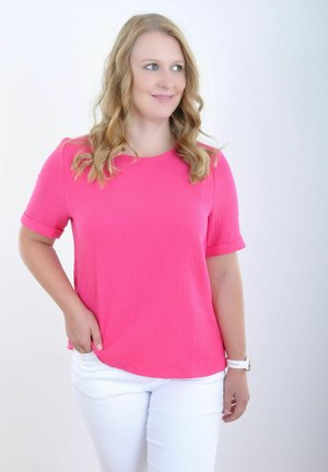 Felroze, getextureerde gebreide top met korte mouwen en opgerolde manchetten, gecombineerd met witte skinny broek. Model poseert met hand in de zak.