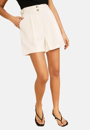 Kvinde iført cremefarvede højtaljede shorts med guldknapper og sorte sandaler med hæl, poserer med den ene hånd i lommen.