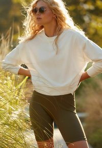 Felpa bianca con polsini a costine, abbinata a pantaloni ciclistici color verde oliva. Il modello indossa occhiali da sole, sullo sfondo di un contesto naturale.