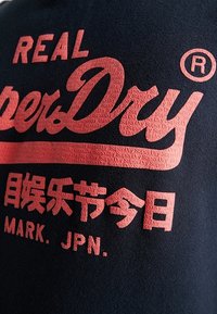 Sudadera negra de algodón con letras rojas en relieve y textura, que muestran "REAL SuperDry" y branding adicional en chino e inglés.