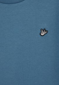 Blå bomullströja med ribbad rundhals. Har en vit broderad fågel-logga på vänster sida, vilket ger en subtil detalj till designen.