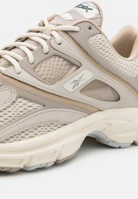 Beige athletic shoe with mesh and suede material, textured detailing, lace-up closure, and a white sole featuring subtle gray accents.