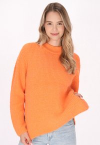 Oranger Strickpullover mit geribbelter Textur, hohem Kragen und überschnittenen Schultern, kombiniert mit hellblauen Jeans.