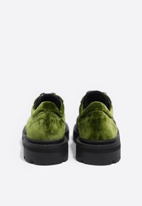 Zapatos de terciopelo verde con una textura suave, presenta una suela gruesa negra y forma de punta redonda. Sin hardware ni patrones visibles.