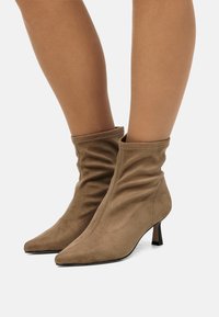 Botas de ante marrón hasta el tobillo con un diseño holgado, punta afilada y un pequeño tacón apilado. Textura suave, costuras mínimas y sin herrajes.
