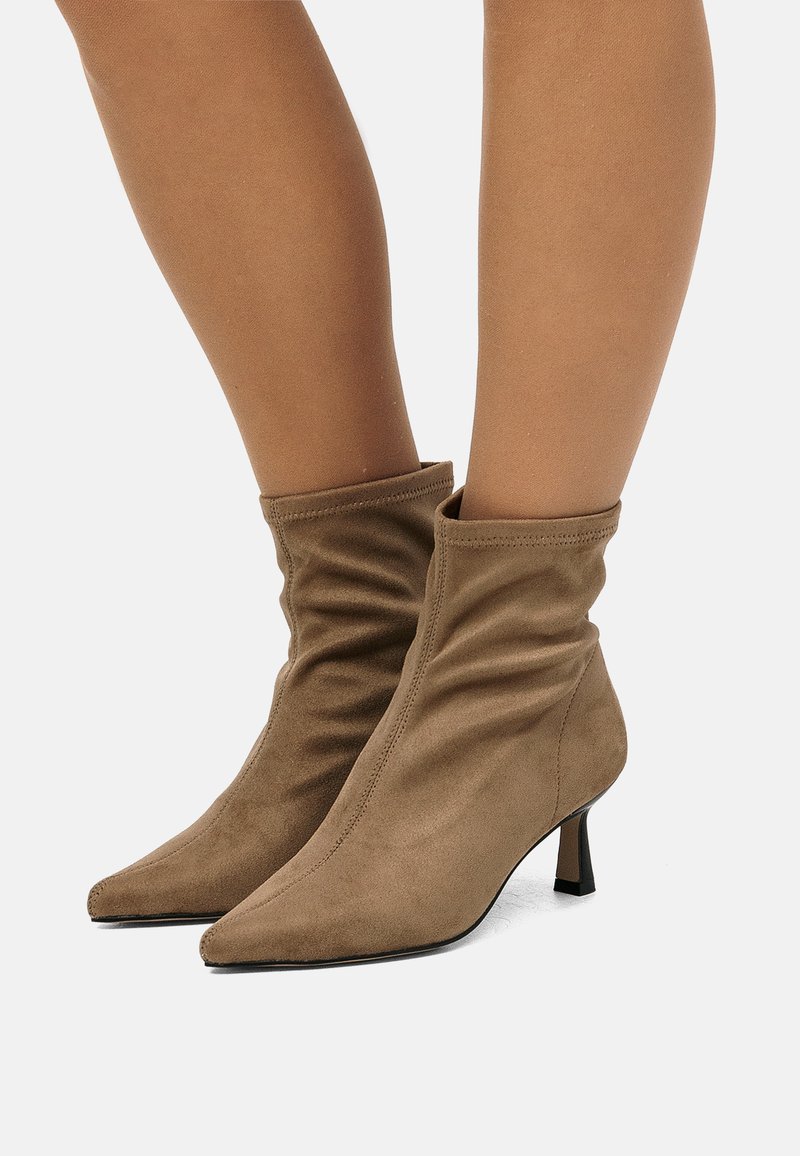 Botas de ante marrón hasta el tobillo con un diseño holgado, punta afilada y un pequeño tacón apilado. Textura suave, costuras mínimas y sin herrajes.