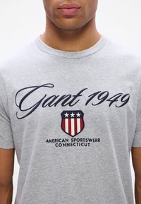 T-shirt de algodão cinza com texto bordado em azul marinho "Gant 1949" e um logótipo de escudo com riscas vermelhas e brancas, rotulado "American Sportswear Connecticut."