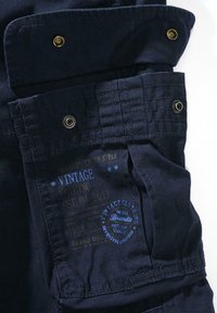 Navyfärgad tygficka med guldsnapsknappar, med blå vintage-tryck och sydda detaljer. Texten inkluderar varumärke och etableringsår.