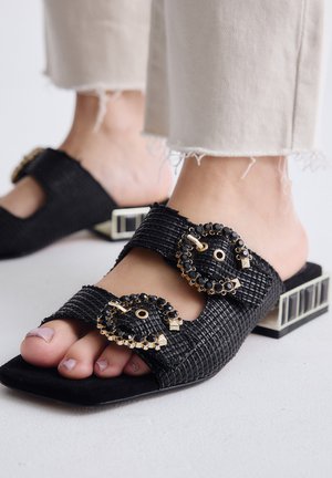 Pies con sandalias tejidas negras de punta abierta, con hebillas decorativas con joyas y tacones bajos de bloque, combinadas con pantalones claros con dobladillo deshilachado.