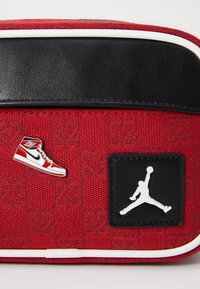 Rote Textiltasche mit schwarzem Lederoberteil, ausgestattet mit weißen Akzenten, einem kleinen roten Sneakers-Anstecker und einem schwarzen Jordan-Logo-Patch.