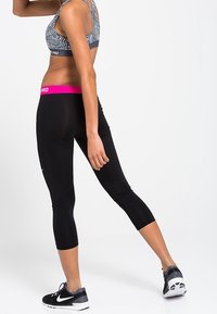Žena oblečená v sportovním brá z a černých zkrácených legínách s růžovým pasem, pózující v černých botách Nike na bílém pozadí.
