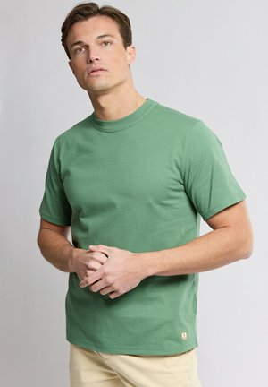 Homme portant un t-shirt vert uni à manches courtes et un pantalon beige, debout les mains jointes devant un fond gris clair.
