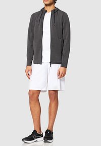 Sweat à zip gris avec capuche, porté sur un t-shirt blanc et un short blanc. Des baskets noires complètent la tenue. Texture lisse, couleurs neutres.