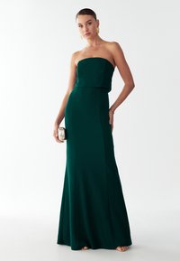 Szmaragdowo-zielona, bezramiowa suknia z dopasowanym gorsetem i rozkloszowanym dołem, wykonana z gładkiego materiału. Modelka trzyma złotą torebkę, nosi złote kolczyki.