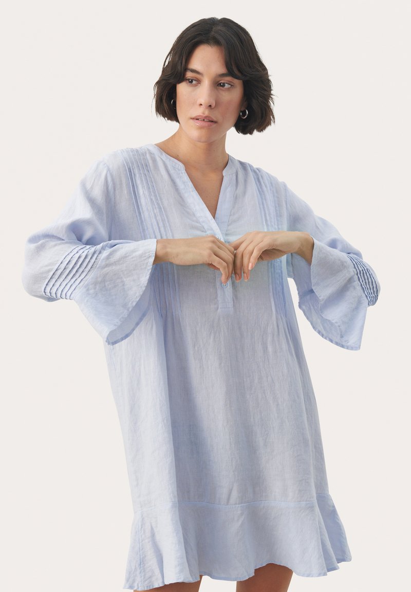 Rochie din in bleu deschis, cu decolteu în V, detalii plisate și mâneci evazate. Prezintă un tiv ondulat și accente subtile texturate.