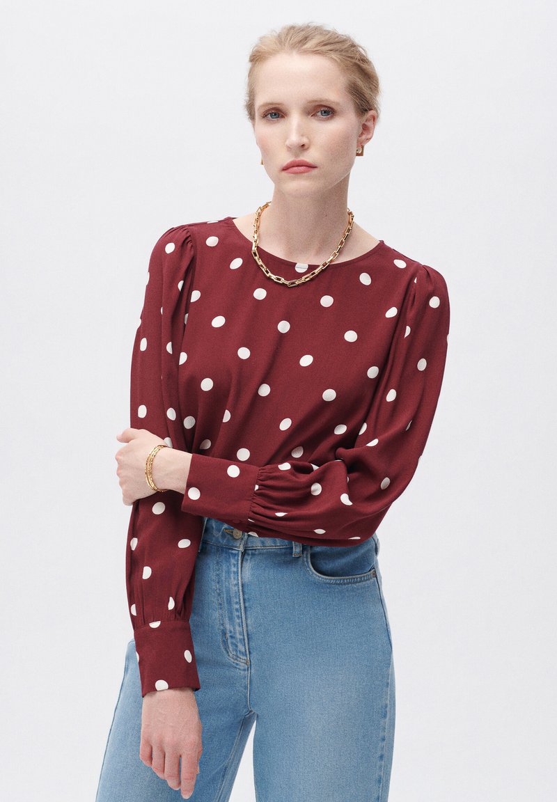 Bordeaux blouse met witte stippen, lange pofmouwen en een ronde hals. Gepaard met lichtblauwe jeans en gouden kettingaccessoires.