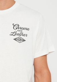 Camiseta blanca de manga corta con un logo impreso en negro que lleva el texto "Chrome and Leather" y "Speed Replay Shop" en forma de emblema.