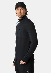 Mann trägt eine schwarze Jacke zum Reißverschluss, schwarze Hose und eine schwarze Baseballkappe, steht und lächelt vor einem schlichten Hintergrund.