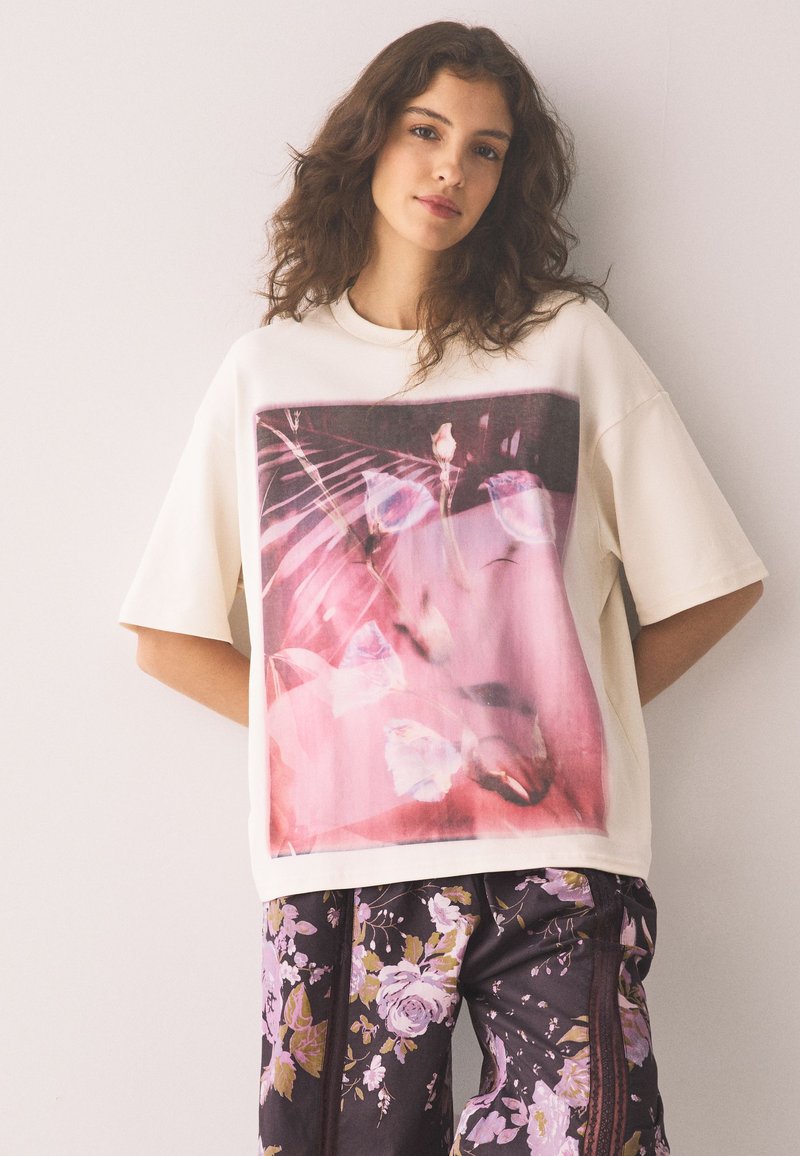 Jeune femme aux cheveux ondulés porte un t-shirt blanc oversize avec un imprimé abstrait rose et un pantalon noir avec un motif floral violet.