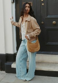 Barna velúr bombázó dzseki fehér csipkés top fölött, széles, világoskék farmernadrág és barna válltáska. Mintás cipők, mögötte egy fekete ajtó.