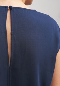 Vue arrière d'une personne portant une robe bleu marine avec une ouverture en clé dans le dos, fermée par un petit bouton recouvert de tissu.