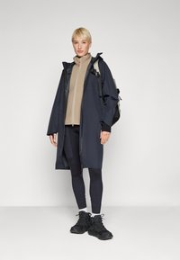 Navy long raincoat with hood over beige fleece jacket, paired with dark leggings and black sneakers. Features zip closure and backpack.