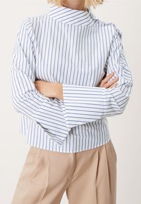 Vit topp med blå vertikala ränder, hög krage och utsvängda ärmar. Har knappdetaljer på axeln och en rak fåll.