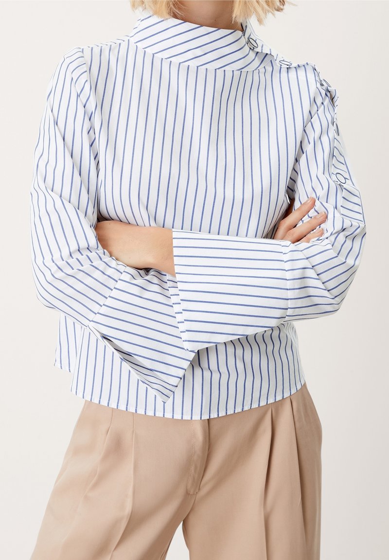 Vit topp med blå vertikala ränder, hög krage och utsvängda ärmar. Har knappdetaljer på axeln och en rak fåll.
