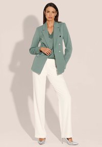 Dubbelzijdige blazer in gedempt teal, met scherpe revers en zilveren knopen, gecombineerd met vloeiende witte broeken en metallic hakken.