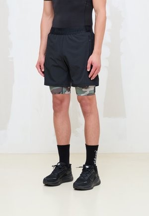 Hombre con pantalones cortos deportivos negros sobre leggings de compresión con estampado de camuflaje, calcetines negros con texto blanco y zapatillas Nike negras.