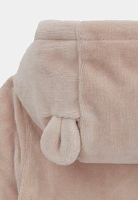 Tejido de corduroy suave en color beige con una superficie texturizada. Presenta un borde enrollado y un detalle anudado en la costura.