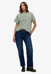 Camiseta de algodón verde claro con "SUPERDAY with NY Ess." impreso en verde más oscuro. Combinada con jeans de pierna recta azul oscuro y zapatos negros.