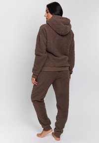 Brun fleece loungewear sæt med hætte sweatshirt og nederdele med ribkant, der har et tekstureret, fluffy materiale og kontrastfarvet brune detaljer.