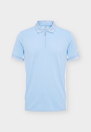 Hombre con una camisa polo de manga corta azul claro, pantalones cortos negros con cinturón negro, calcetines blancos y zapatillas blancas, de pie mirando hacia adelante.