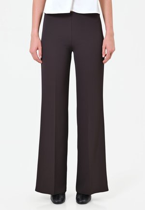 Vrouw draagt zwarte broek met wijde pijpen, zwarte schoenen en een wit topje, staand voor een effen lichte achtergrond.