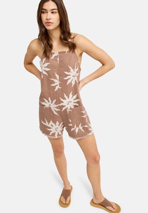 Frau steht mit Händen in der Hüfte, trägt braunen ärmellosen Overall mit weißen Sonnentatzen und braune Sandalen.