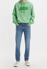 Personne portant un sweat à capuche Sequoia vert clair, un jean bleu et des baskets blanches, debout les mains dans les poches devant un fond uni.