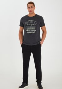Graues Graphic-T-Shirt mit vintage Textdesign, kombiniert mit schwarzen Jogginghosen und schwarzen Sneakers. Lässiger Schnitt, glatter Stoff.