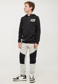 Felpa nera con zip e stampa bianca sul petto, abbinata a pantaloni della tuta grigi con pannelli laterali neri. Sneakers con dettagli in bianco e nero.