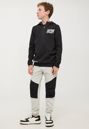 Felpa nera con zip e stampa bianca sul petto, abbinata a pantaloni della tuta grigi con pannelli laterali neri. Sneakers con dettagli in bianco e nero.