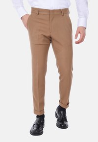 Pantaloni su misura marrone chiaro con una superficie liscia, vestibilità slim, pieghe frontali, tasche laterali e orli risvoltati, abbinati a mocassini neri.