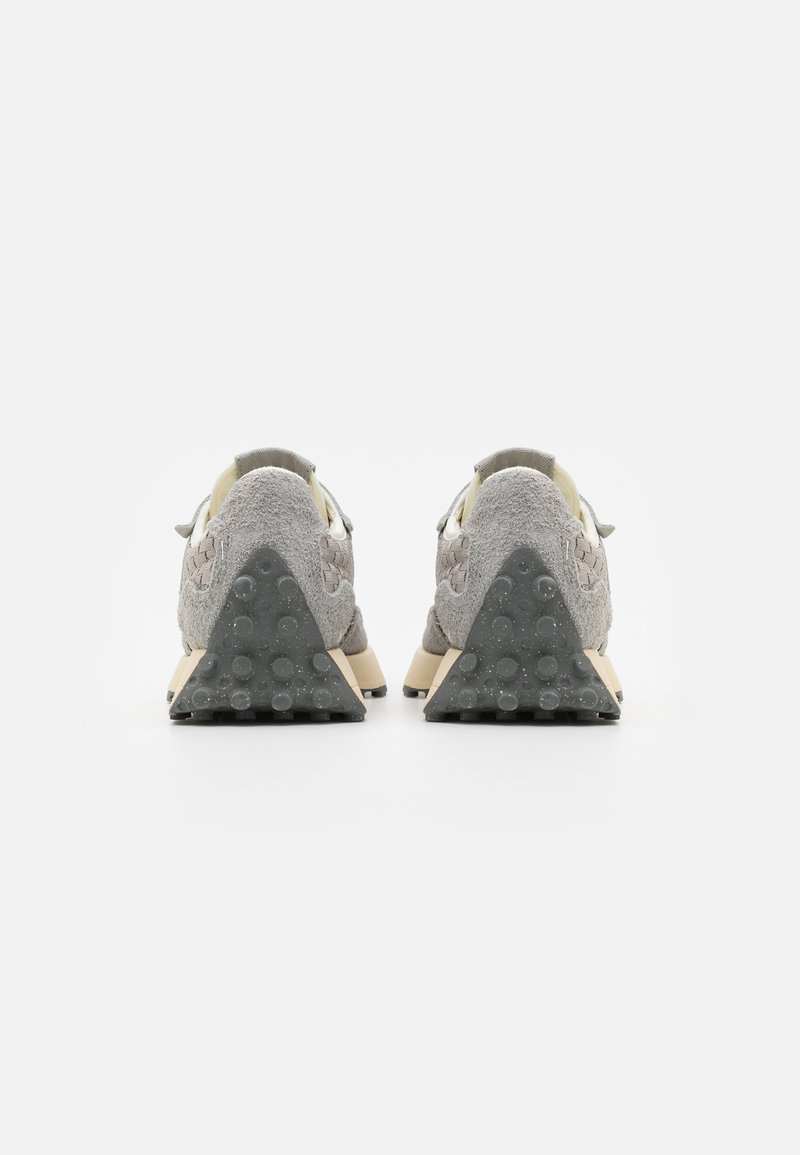 Un par de zapatillas de ante gris con contrafuertes de goma texturizada en el talón y detalles laterales trenzados, mostradas desde atrás sobre un fondo blanco.