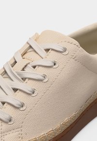 Κοντινό πλάνο μπεζ πάνινο αθλητικό παπούτσι με λευκά κορδόνια και υφασμάτινη λεπτομέρεια πάνω από καφέ λαστιχένια σόλα σε λευκό φόντο.