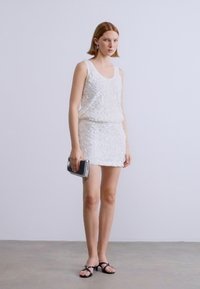 Λευκό αμάνικο φόρεμα με υφή και στρωματοποιημένη σχεδίαση· μίνι μήκος. Συνδυασμένο με μαύρα σανδάλια και ασημένιο τσαντάκι. Απλό φόντο.