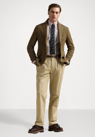 Blazer en tweed marron, chemise rayée, cravate à motifs, pantalon beige clair, chaussures à lacets marron. Le blazer a un col à revers et un bouton unique.