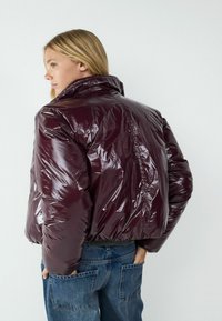 Burgunderfarbene, glänzende Puffjacke mit hohem Kragen, tailliert und mit glatter Oberfläche. Kombiniert mit blauen Jeans.
