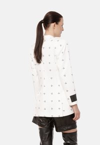 Biała marynarka z czarnymi abstrakcyjnymi wzorami, długie rękawy i czarny akcent na mankietach. Projekt wyróżnia się dopasowanym tyłem z rozcięciem.