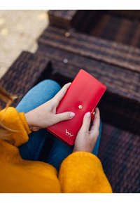 Red leather wallet with a snap closure, featuring gold hardware and a logo. Held in hands, against a textured, dark wooden surface.